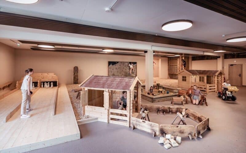 Die Kinder spielen in einem Innenbereich mit Holztiergehegen, kleinen Häusern und einer Bowlingbahn, an der auch ein Kind im Rollstuhl teilnimmt.
