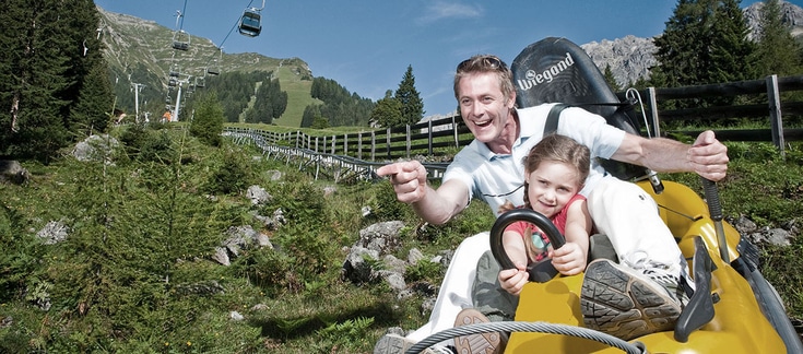 Ein Mann und ein junges Mädchen fahren mit einem gelben Alpine Coaster einen grasbewachsenen Berghang hinunter, mit Sesselliften und Kiefern im Hintergrund.