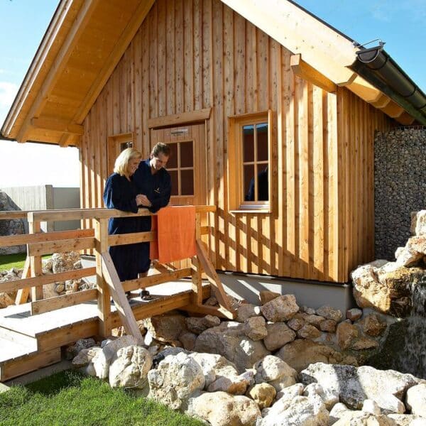 Zwei Personen stehen auf einer Holzrampe vor einer kleinen Holzhütte und blicken auf einen Steingarten mit einem Wasserfall.