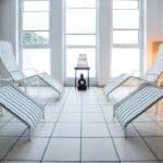 Vier weiße Loungesessel mit blauen Streifen stehen in einem hellen, gefliesten Raum mit großen Fenstern und dekorativer Wandkunst. In der Mitte steht ein kleiner Tisch mit einer Lampe.