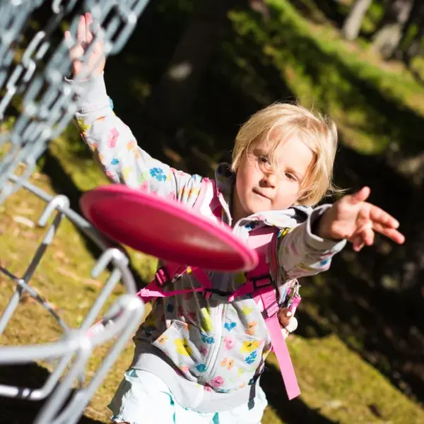 Ein kleines Kind in einer gemusterten Jacke wirft eine rosafarbene Scheibe auf einen Disc-Golf-Korb aus Metall in einem sonnenbeschienenen Waldgebiet.
