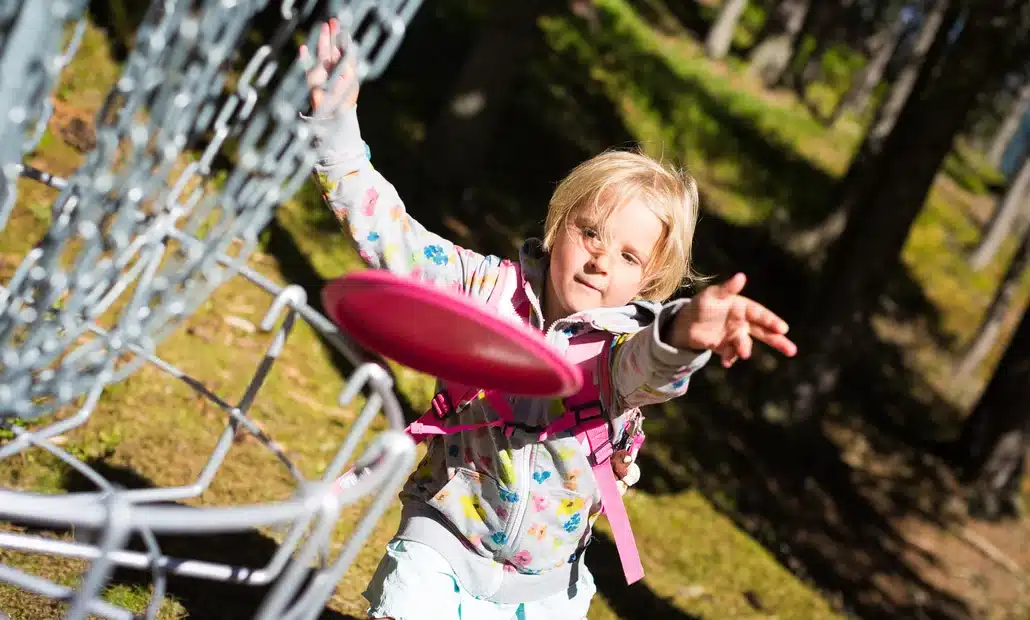 Ein kleines Kind in einer gemusterten Jacke wirft eine rosafarbene Scheibe auf einen Disc-Golf-Korb aus Metall in einem sonnenbeschienenen Waldgebiet.