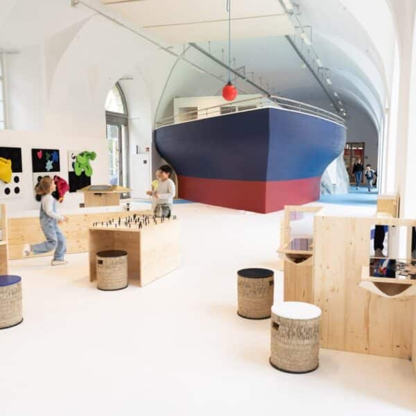 Kinder spielen in einem geräumigen, modernen Raum mit Holzmöbeln und einer großen Bootskonstruktion im Hintergrund.
