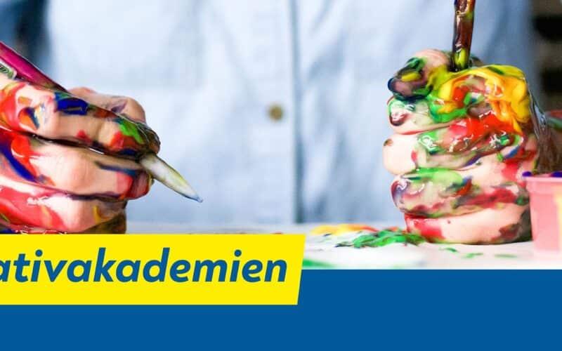 Eine Person mit farbverschmierten Händen hält Pinsel über einen Tisch mit bunten Farben. Auf einem gelb-blauen Banner steht der Text „Kreativakademien“.