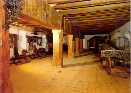 Ein historischer Raum mit Holzbalken und Säulen, in dem alte Möbel, dekorative Elemente und alte Geräte an den Wänden ausgestellt sind.