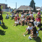 Eine Gruppe von Kindern und zwei Erwachsene sitzen und stehen an einem sonnigen Tag auf einem Grashügel im Freien und wirken entspannt und in ein Gespräch oder ein Spiel vertieft.