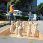 Ein Kind wirft eine Kugel, um große hölzerne Bowlingkegel auf einer hölzernen Außenbahn umzuwerfen, während ein Erwachsener in der Nähe steht.