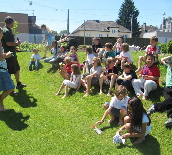 Eine Gruppe von Kindern sitzt und steht an einem sonnigen Tag auf einem Grashügel im Freien, im Hintergrund sind Häuser und Bäume zu sehen.