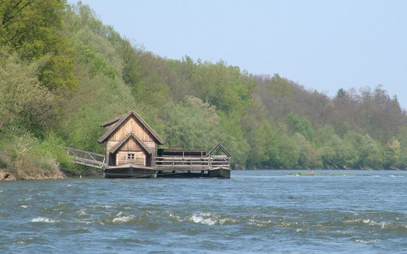Ein hölzernes Bootshaus steht am Rande eines Flusses, umgeben von dichten grünen Bäumen unter einem klaren Himmel.