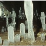 Mehrere unterschiedlich hohe Eisstalagmiten ragen aus dem Höhlenboden, während im Hintergrund Licht aus der Höhlenöffnung einfällt.