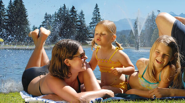 Eine vierköpfige Familie entspannt sich an einem sonnigen Tag auf einer Decke an einem See, im Hintergrund sind Bäume, Berge und rote Sonnenschirme zu sehen.