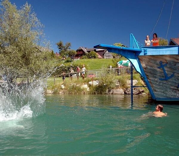 Die Leute schwimmen und sehen zu, wie jemand von einer blauen Holzkonstruktion in Form eines Schiffes in einen See springt und dabei einen großen Sprung macht.