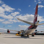 Ein großes Verkehrsflugzeug mit rotem Leitwerk steht bei teilweise bewölktem Himmel auf der Rollbahn, in der Nähe befinden sich Fahrzeuge des Bodenservice.