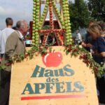 Menschen stehen bei einer Veranstaltung im Freien neben einem Holzschild mit der Aufschrift "Haus des Apfels", im Hintergrund eine Apfelauslage aus gestapelten Äpfeln.