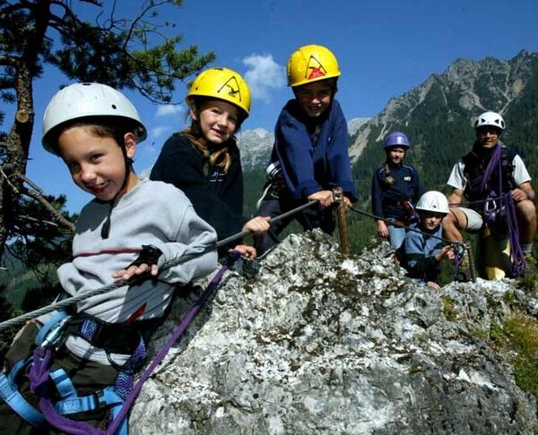 Eine Gruppe von Kindern und ein Erwachsener, die alle Helme und Klettergurte tragen, klettern auf einen Felsen in einer bergigen Umgebung.