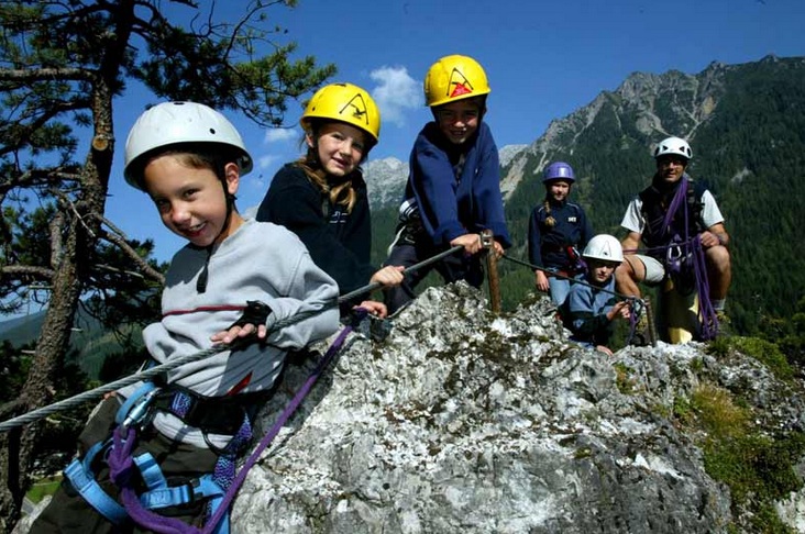 Eine Gruppe von Kindern und ein Erwachsener, die alle Helme und Klettergurte tragen, klettern auf einen Felsen in einer bergigen Umgebung.