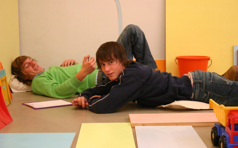 Zwei junge Menschen liegen in einem Raum mit bunten Oberflächen auf dem Boden, einer lächelt in die Kamera, der andere schreibt auf einem Klemmbrett. In der Nähe sind ein Spielzeuglastwagen und ein orangefarbener Eimer zu sehen.