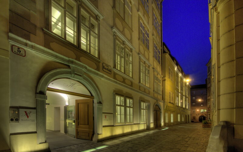 Eine schmale Kopfsteinpflasterstraße mit historischen Gebäuden unter einem tiefblauen Abendhimmel; ein Gebäude hat ein offenes Bogentor, dessen Eingang von Lichtern beleuchtet wird.