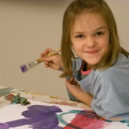 Ein junges Mädchen lächelt, während es mit einem Pinsel an einem Tisch malt und bunte Kunstwerke auf Papier schafft.