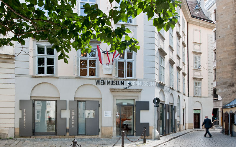 Straßenansicht des Wien Museum Uhrenmuseums, ein weißes Gebäude mit großen Fenstern, Beschilderung und mehreren Fahnen über dem Eingang in einer Kopfsteinpflasterstraße.