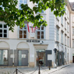 Außenansicht des Wien Museum Uhrenmuseums, mit großen Fenstern, Beschilderung und ein paar Menschen, die in der Nähe auf einer Kopfsteinpflasterstraße spazieren gehen.