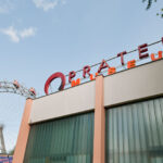 Ein Gebäude mit der Aufschrift "Pratermuseum" neben einem großen Riesenrad, das bei klarem Himmel teilweise hinter Bäumen sichtbar ist.