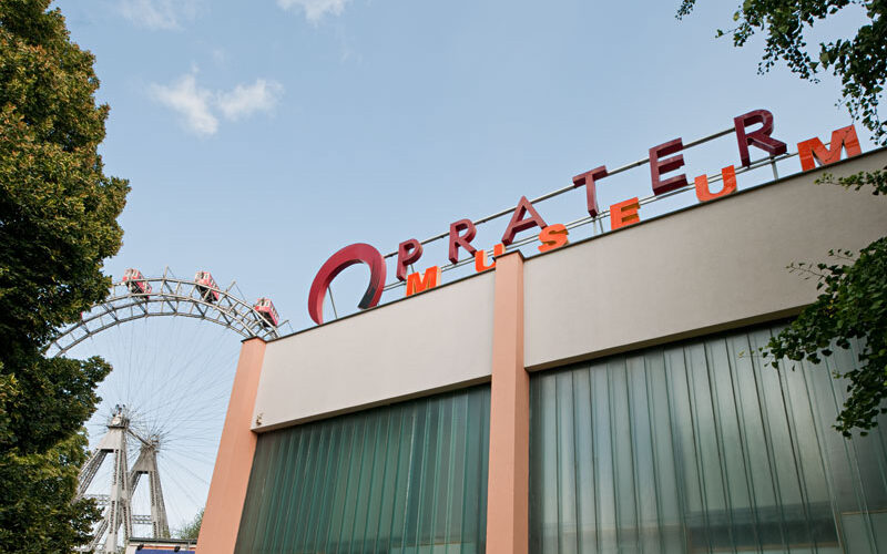 Ein Gebäude mit der Aufschrift "Pratermuseum" neben einem großen Riesenrad, das bei klarem Himmel teilweise hinter Bäumen sichtbar ist.