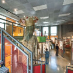 Innenansicht einer Museumsausstellung mit Schautafeln, gerahmten Gegenständen und einer großen Figur mit einer Blumenkrone und einem Korb auf dem Kopf neben einer Treppe.