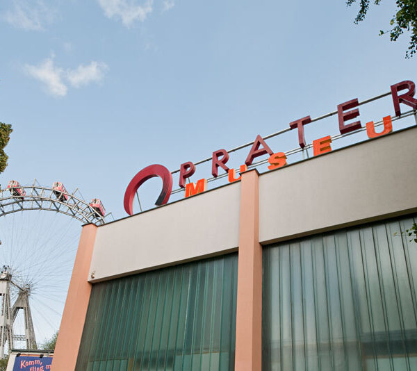 Gebäude des Pratermuseums mit Teilansicht des Wiener Riesenrads im Hintergrund, bei klarem Himmel und umgeben von Bäumen.