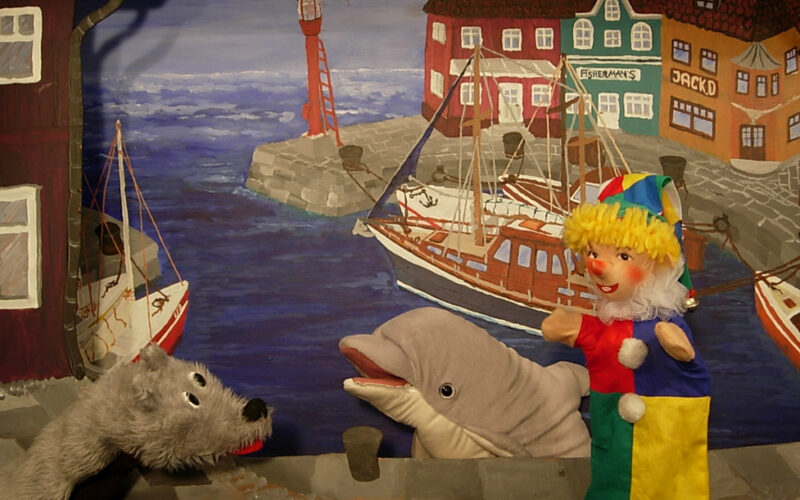 Drei Puppen - ein grauer Wolf, ein Delfin und ein Clown - treten vor einer gemalten Kulisse auf, die einen Hafen mit Booten und bunten Gebäuden zeigt.