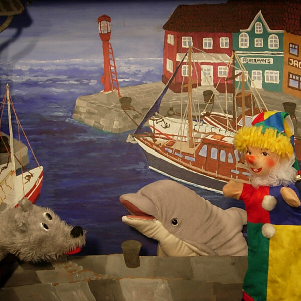 Drei Puppen - ein grauer Hund, ein Delfin und ein Clown - stehen vor einem gemalten Hintergrund, der einen Hafen mit Booten und bunten Gebäuden zeigt.