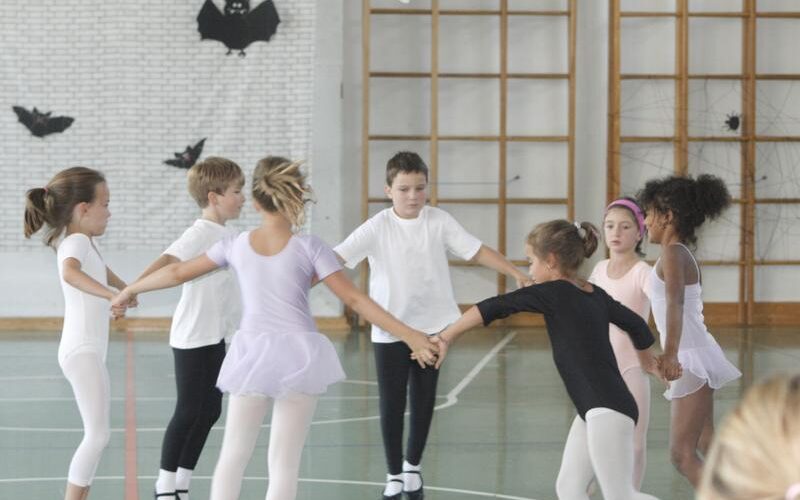 Eine Gruppe kleiner Kinder in Ballettkleidung hält sich in einer mit schwarzen Papierfledermäusen an den Wänden dekorierten Turnhalle im Kreis.