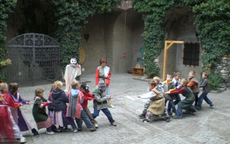 Eine Gruppe kostümierter Kinder spielt Tauziehen in einem gepflasterten Innenhof. Ein Erwachsener beaufsichtigt die Kinder und in der Nähe steht eine Person mit einer weißen Maske und einem Umhang.