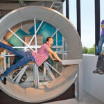 Ein Kind dreht sich in einem großen menschlichen Hamsterrad, während zwei andere Kinder in einem hell erleuchteten Innenraum von einem nahen Sims aus zusehen.