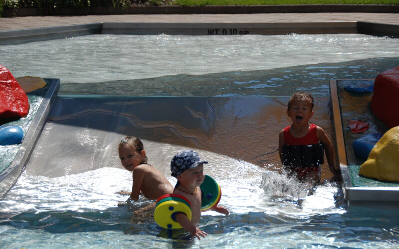 Drei kleine Kinder spielen im seichten Wasser eines Planschbeckens mit kleinen bunten Strukturen und Wasser, das über eine Rampe fließt.