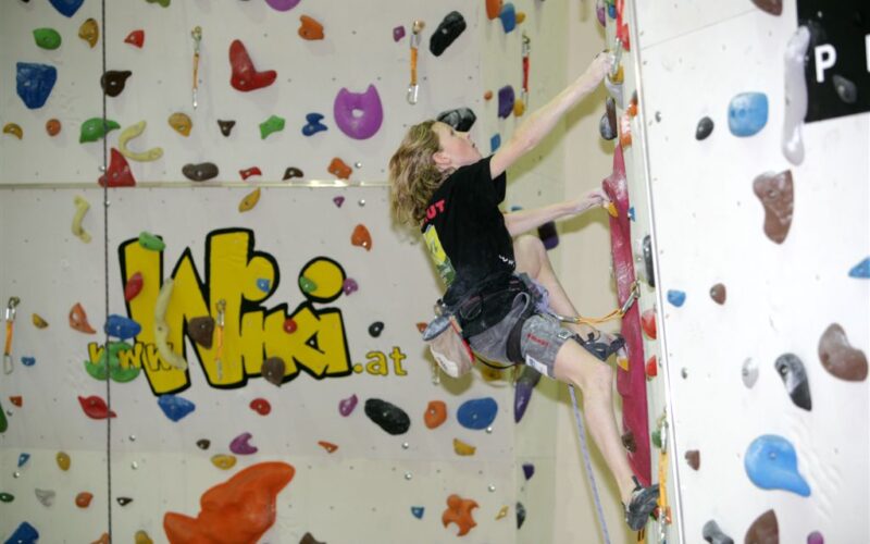 Eine Person in Kletterausrüstung besteigt eine Indoor-Kletterwand mit bunten Griffen; im Hintergrund ist ein großes "WIKI"-Logo zu sehen.