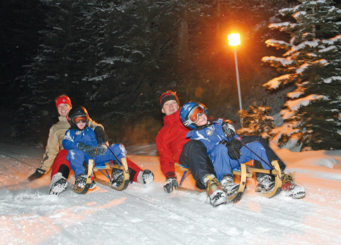 Zwei Erwachsene und zwei Kinder in Winterkleidung fahren nachts auf einem verschneiten Weg Schlitten, mit Bäumen und einer Straßenlaterne im Hintergrund.