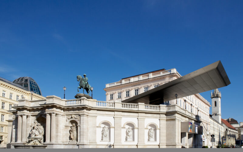 Ein großes Reiterstandbild steht auf einem klassischen Steinbau mit Bögen und Skulpturen neben einem modernen, kantigen Dach unter einem klaren blauen Himmel.