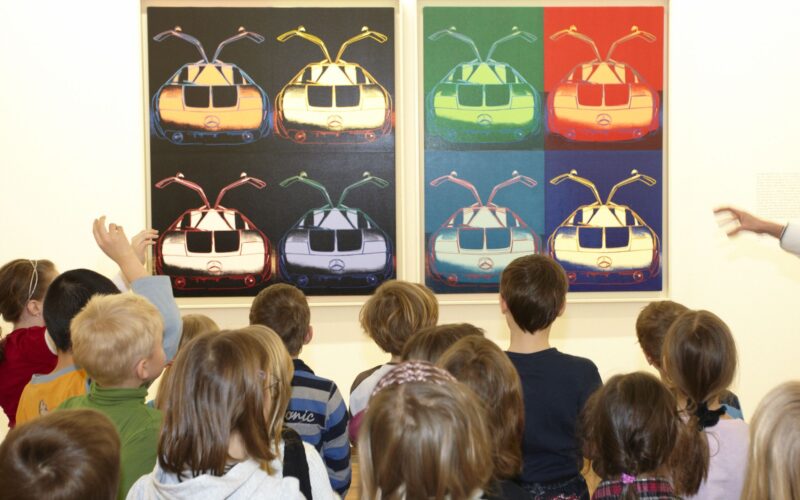 Eine Gruppe von Kindern steht vor zwei farbenfrohen Kunstwerken, die jeweils mehrere stilisierte Bilder einer Straßenbahn im Pop-Art-Stil zeigen.