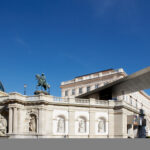 Ein modernes Metalldach erstreckt sich über einer historischen Reiterstatue und einem Steindenkmal mit klassischen Skulpturen, die vor der Kulisse städtischer Gebäude unter einem klaren blauen Himmel stehen.