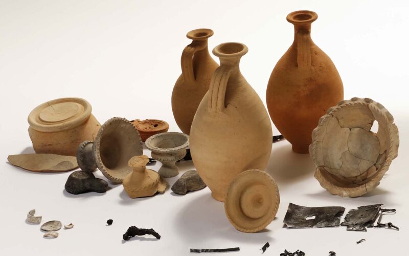 Eine Sammlung von antiken Keramikgefäßen, Schalen, Deckeln, Münzen, Metallgegenständen und Keramikfragmenten, die auf einem schlichten weißen Hintergrund angeordnet sind.