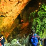 Menschen mit Rucksäcken erkunden bei künstlicher Beleuchtung eine große Höhle mit zerklüfteten, teilweise mit grünem Moos bewachsenen Felsformationen.