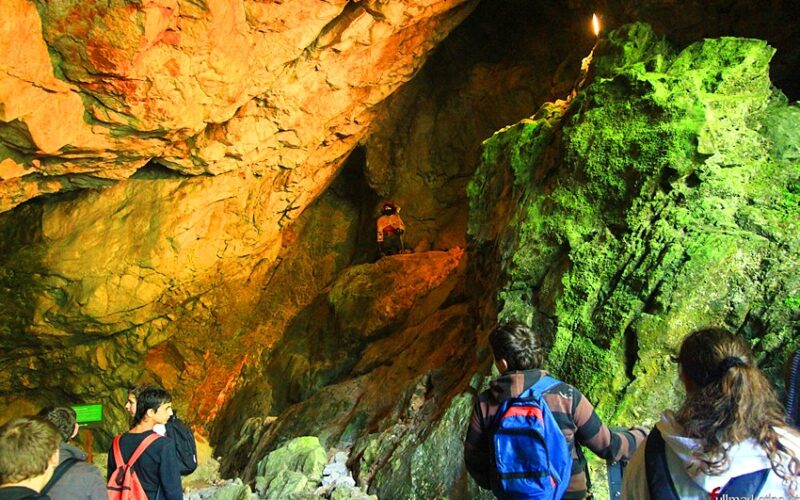 Menschen mit Rucksäcken erkunden bei künstlicher Beleuchtung eine große Höhle mit zerklüfteten, teilweise mit grünem Moos bewachsenen Felsformationen.