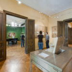 Menschen, die Kunstwerke in einem geräumigen, gut beleuchteten Museumsraum mit Holzböden, großen Türen und Kunstwerken an den Wänden und in einer Vitrine betrachten.