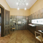 Ein Museumsraum mit Holzböden und -wänden, Vitrinen mit kleinen Skulpturen, gerahmten Kunstwerken, zwei Holzstühlen und Deckenbeleuchtung.