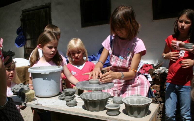 Eine Gruppe von Kindern steht im Freien um einen Tisch herum, während ein Mädchen auf einer Töpferscheibe Ton formt, während auf dem Tisch Tonobjekte und Formen ausgestellt sind.