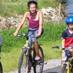 Vier Kinder mit Helmen fahren an einem sonnigen Tag auf einem gepflasterten Weg neben Gras und Steinen Fahrrad.