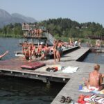 Die Menschen sonnen sich und schwimmen auf hölzernen Stegen an einem See mit Sprungtürmen, umgeben von Bäumen und Bergen im Hintergrund.