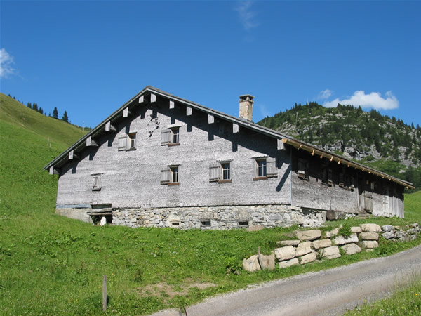 Ein großes, rustikales Bauernhaus aus Holz mit einem Fundament aus Stein steht auf einem grasbewachsenen Hügel unter einem blauen Himmel mit vereinzelten Wolken. Ein Schotterweg führt neben dem Gebäude entlang.