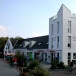 Ein weißes, modernes Gästehaus mit eckigen Dächern und großen Fenstern; draußen stehen Menschen mit Fahrrädern, und im Erdgeschoss sind ein paar Geschäfte zu sehen.